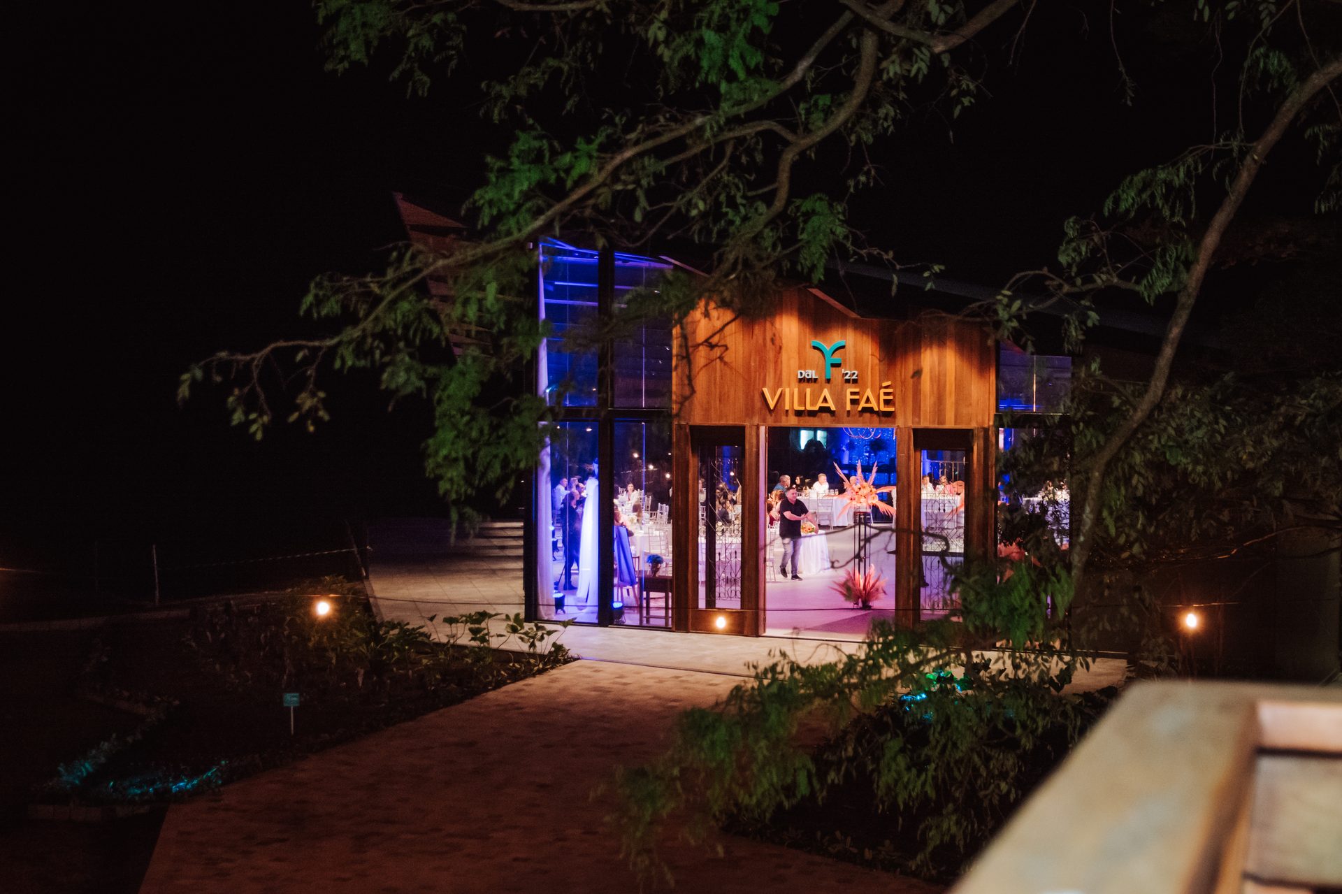 Fachada externa noturna da Villa Faé com jardim iluminado